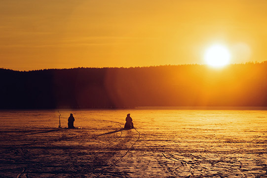 Photo shows two men ice fishing on a lake ice and the Sun shining just above the horizon in the daytime.