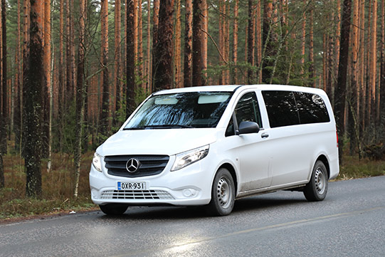 In the photo a white Mercedes mini bus drives vie a beautiful pine forest road.